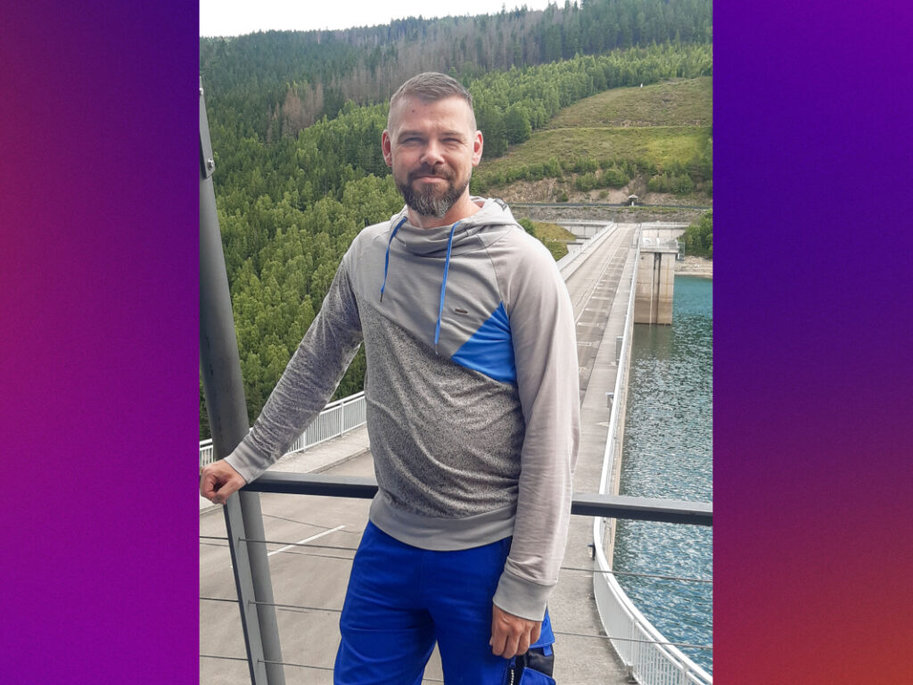 Ein Mann mit sportlicher Kleidung steht vor dem Damm einer Talsperre. Im Hintergrund sieht man Wald.