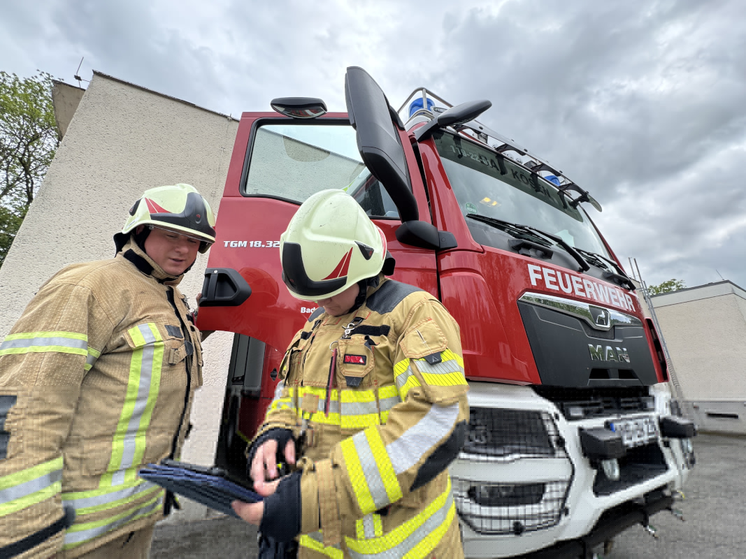 Zwei Feuerwehrleute in Montur stehen vor einem Feuerwehrfahrzeug und schauen auf den Bildschirm eines Tablets.